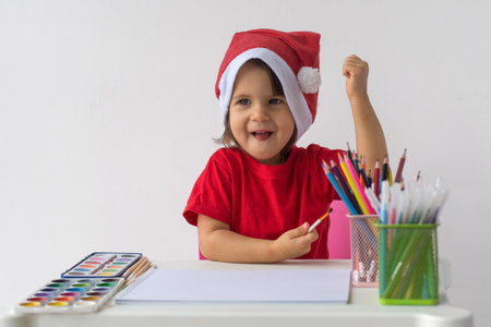 A happy little girl in a Santa hat and red t-shirt sits at a table with art supplies, ready to create festive drawings. She smiles joyfully, celebrating the holiday season. Concept of Christmas creativityの写真素材