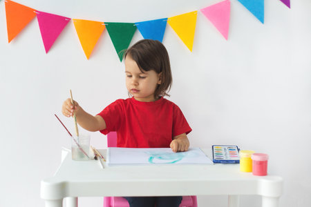 girl 2-3 years child in a red t-shirt is painting with watercolors at a white table, dipping a brush into a glass of water, with colorful banners in the backgroundの写真素材
