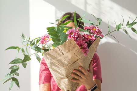 Woman in pink shirt holding bouquet of chrysanthemums and greenery wrapped in vintage newspaper, hiding her face behind flowers in sunlight, concept of freshness, femininity and natural beautyの写真素材