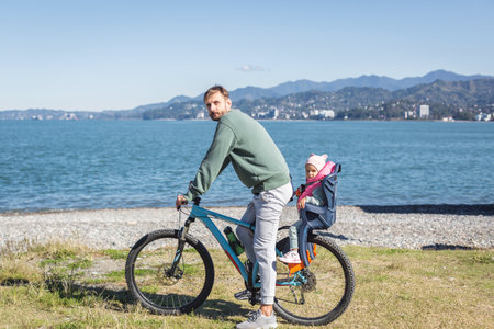 Father riding mountain bike with his little daughter in child seat by the sea on sunny day, both wearing casual clothes, mountains cityscape in background, concept of family travel outdoor recreationの写真素材