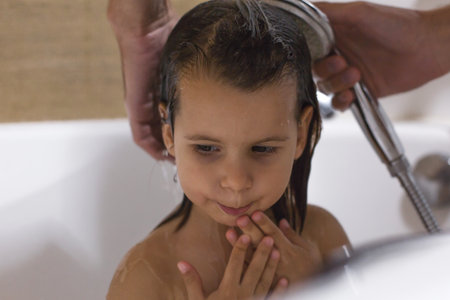 Little child taking a bath while an adult rinses their hair with a shower head, showing care, hygiene, and family bonding during daily routine in a cozy home bathroomの写真素材