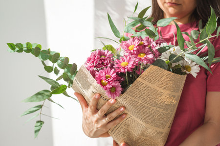 woman in pink shirt gently holds bright bouquet of pink white flowers mixed with lush green leaves, wrapped in old newspaper. Sunlight soft petals textures, creating a cozy, nostalgic moodの写真素材