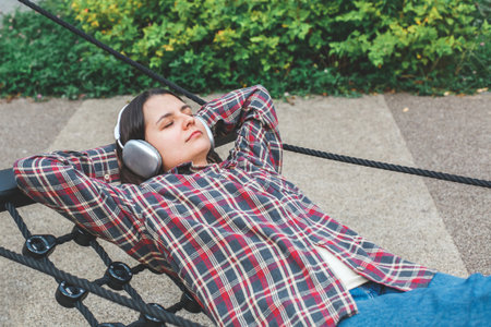 Young woman in checkered shirt relaxing outdoors on rope swing with wireless headphones, eyes closed, concept of leisure, music, mindfulness and modern lifestyle balanceの写真素材