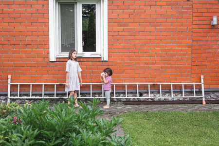 Two children standing near a red brick house wall with a ladder lying on the ground, showing everyday family life, communication, and childhood moments in a suburban home environmentの写真素材