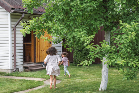 Two barefoot children running across a green garden toward a small wooden house on a sunny day,...