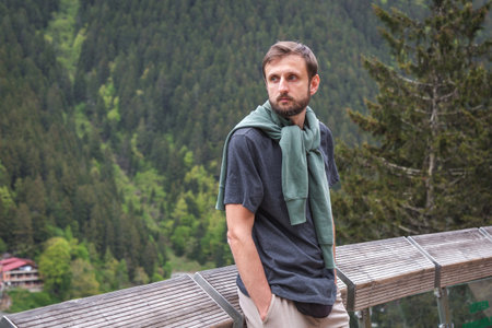 Bearded man in a dark t-shirt with a green sweatshirt over his shoulders stands on a wooden terrace...