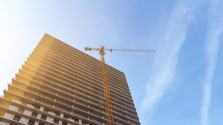 Highrise building under construction with a tall yellow tower crane lifting materials, illuminated by warm sunlight against a clear blue sky, symbolizing urban development and modern architectureの写真素材