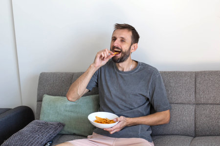 Man relaxing on the couch at home, watching TV and eating snacks. Casual evening routine, leisure time, enjoying chips while sitting comfortably in the living room.の写真素材