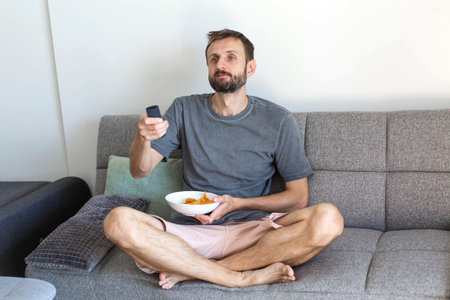 Man sitting on the couch, holding a remote control and turning on the TV while eating snacks. Relaxing at home, watching television and enjoying leisure time in a comfortable living room.の写真素材