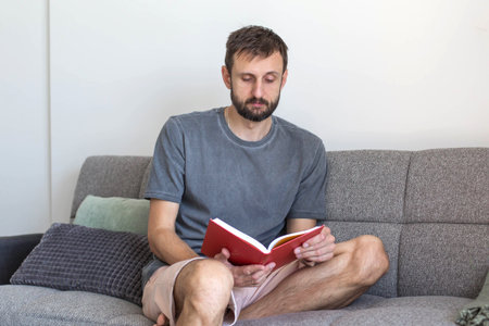 Man sitting on a sofa and reading a book in a bright living room. Relaxed home atmosphere, leisure time, learning and peaceful daily routine in a comfortable modern interior.の写真素材