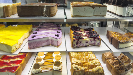 Assorted cheesecake slices displayed in a refrigerated bakery showcase. Variety of colorful cakes...
