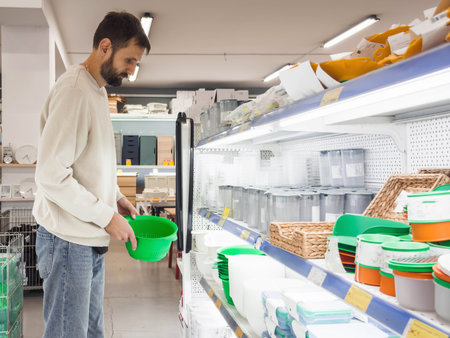 Man shopping in a household goods store, examining a green plastic kitchen bowl among shelves with containers and storage items. Bright lighting, clean interior and everyday shopping atmosphere.の写真素材