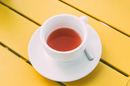 A white cup of hot herbal or fruit tea sits on a bright yellow table, creating a clean, minimalistic, and refreshing composition with vibrant contrasting colors.の写真素材