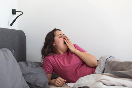 A sleepy woman yawns widely while waking up in bed, covering her mouth with her hand. The candid morning scene shows tiredness, slow awakening, and the natural struggle to start the day.の写真素材