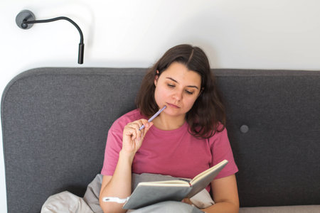 A woman sits in bed thoughtfully holding a pen and writing in her notebook, capturing a calm moment...