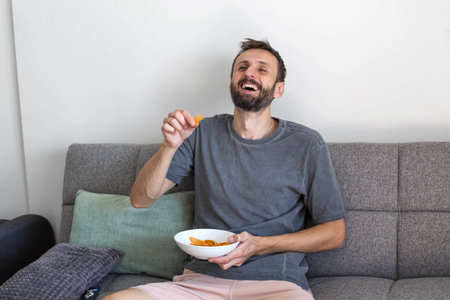 Smiling man sitting on a sofa and eating chips at home. Relaxed leisure time, enjoying a snack and laughing in a cozy living room environment. Casual lifestyle, comfort and positive mood indoors.の写真素材