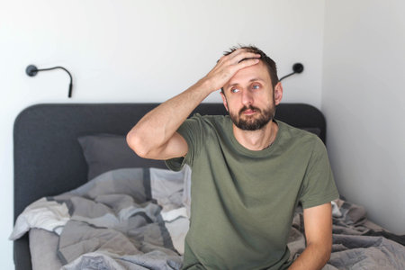 A tired man sitting on an unmade bed in a bright bedroom, holding his head with one hand and looking away with a stressed morning expression, capturing a realistic moment of exhaustion and waking upの写真素材