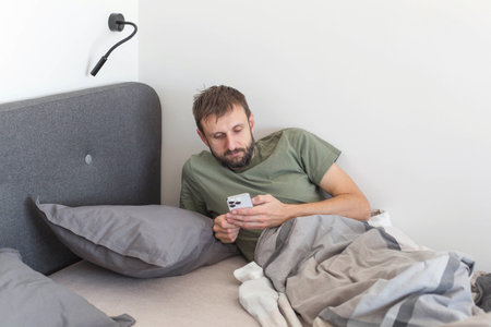 Man lying in bed in modern minimalist bedroom checking smartphone screen under soft natural light...