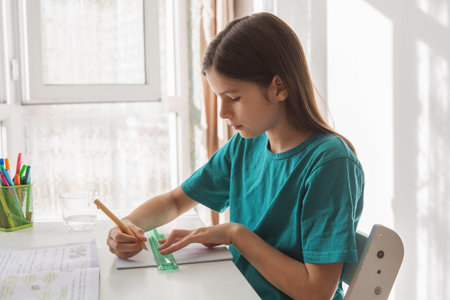 Schoolgirl studying at home at white desk near window writing in notebook with pencil and ruler during daytime, focused child doing homework in bright cozy interior with natural sunlightの写真素材