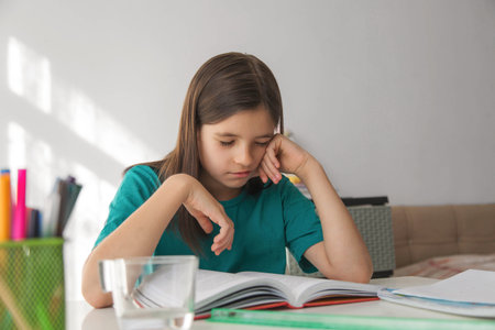 Calm young girl sleeping at a bright home desk with an open textbook, sitting in soft natural light while taking a quiet break from studying in a tidy room with school supplies around herの写真素材