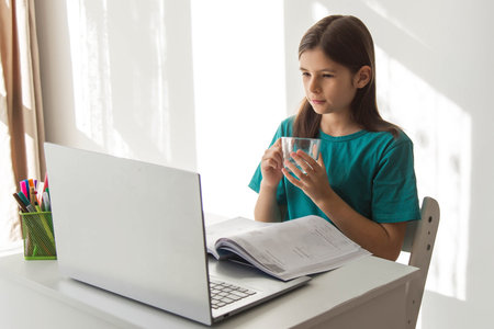 Schoolgirl studying at home with a laptop and open workbook, holding a glass of water while watching the screen, sunlight filling the room and creating a calm atmosphere for focused remote learningの写真素材