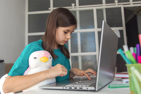 Girl studying at home with a laptop, focused on typing while holding a cute plush penguin, sitting...