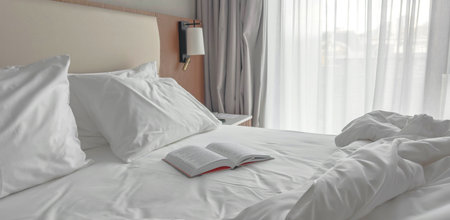 Cozy hotel bedroom with soft white bedding, fluffed pillows an open book resting on bed near large...