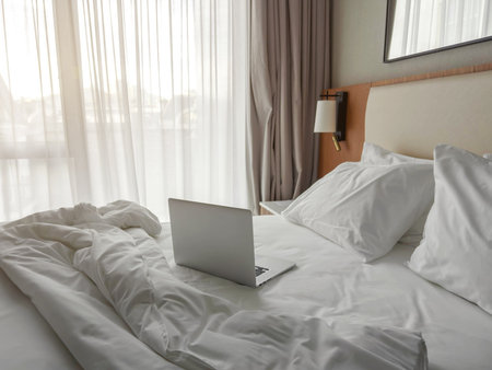 Bright hotel room with soft white bedding, pillows and an open laptop on an unmade bed, creating a cozy atmosphere for remote work, relaxation, travel comfort and modern lifestyleの写真素材