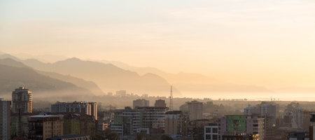 Urban cityscape at sunrise with layered mountains in background, soft morning haze, residential...