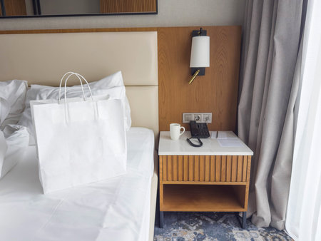 White shopping bag on hotel bed next to bedside table with phone and cup, modern hotel room...