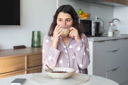 Young woman in pajamas drinking coffee at breakfast table in home kitchen, relaxed morning...