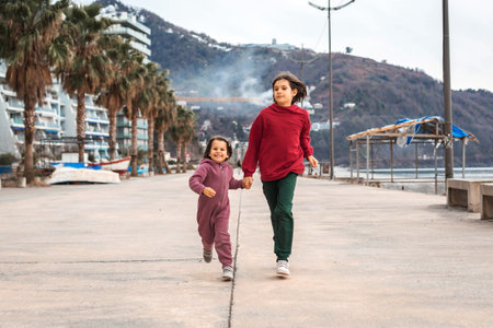Two happy children running hand in hand along the seafront promenade with palm trees and mountains in the background, enjoying sibling bonding time and outdoor activity during vacationの写真素材