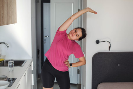 Young woman with dark hair in pink t shirt and black shorts stretching side bend exercise at home...