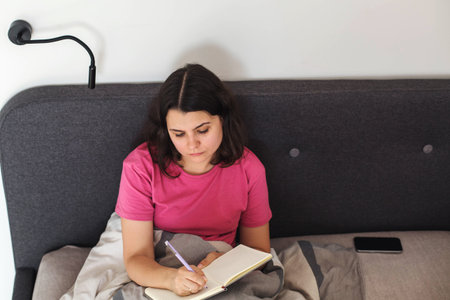 Woman writing in notebook while sitting in bed at home, personal journal, planning thoughts, self...
