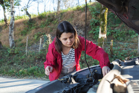 Woman checking car engine under open hood on forest roadside, female driver inspecting vehicle after...