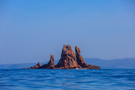A picturesque red stone island sticks out of the blue sea water. An uninhabited rocky island in the...