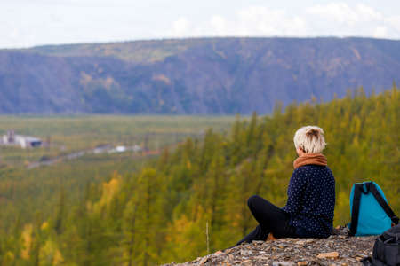 A blond girl sits on a hillock against the backdrop of a beautiful lake with looks into the distance. View from the back.の写真素材