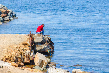 Summer, 2016 - Vladivostok, Russia - Water area of Fedorov Bay in Vladivostok. Lonely fisherman man sits on broken stone pier with fishing rod and looks out to sea.の写真素材