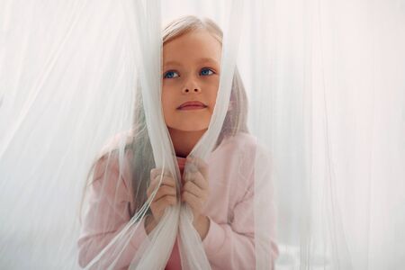 Little beautiful dreamy girl portrait behind window curtain in bedroomの写真素材