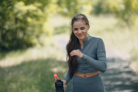 Smiling woman jogger in gray tracksuit drinking bottled water after jogging in park outdoor.の写真素材