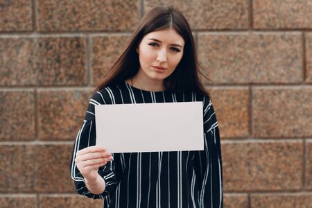 Young woman holds white paper in hand stone background. Girl with white blank template sheet with empty space.の写真素材