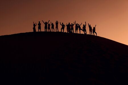 People standing on top of dune in desert with hands up. Silhouettes at sunsetの写真素材