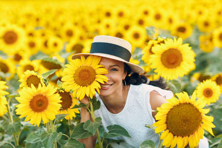 Beautiful smiling young woman in a hat with flower on her eye and face on a field of sunflowersの写真素材