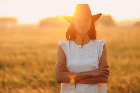 Woman farmer in cowboy hat at agricultural field on sunset with sunflareの写真素材