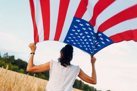 Woman with American flag in wheat field at sunset. 4th of July. Independence Day patriotic holiday.の写真素材