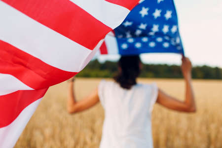 Woman with American flag in wheat field at sunset. 4th of July. Independence Day patriotic holiday.の写真素材