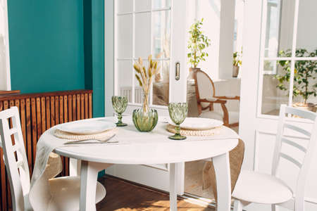 Green glasses and white dishes on the kitchen round table, with green plant on living room in the background.の写真素材