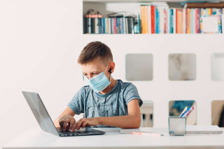 Young boy in headset sitting at table with laptop and medical face mask and preparing to school. Online education concept.の写真素材