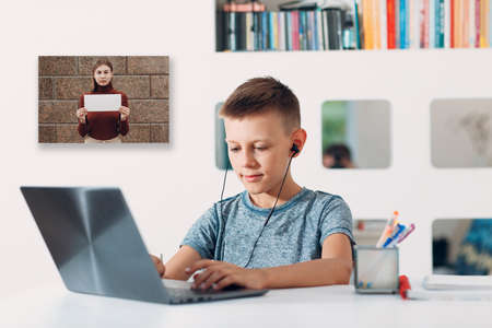 Young boy in headset sitting at table with laptop and preparing to school by teacher with white empty poster copy space on computer screen. Online education concept.の写真素材