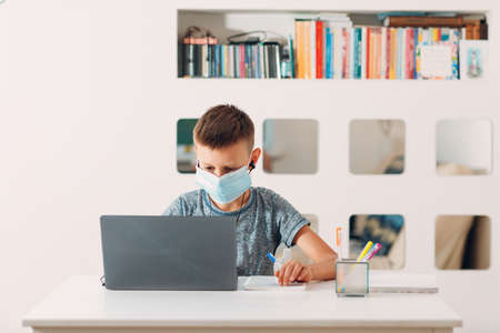 Young boy in headset sitting at table with laptop and medical face mask and preparing to school. Online education concept.の写真素材